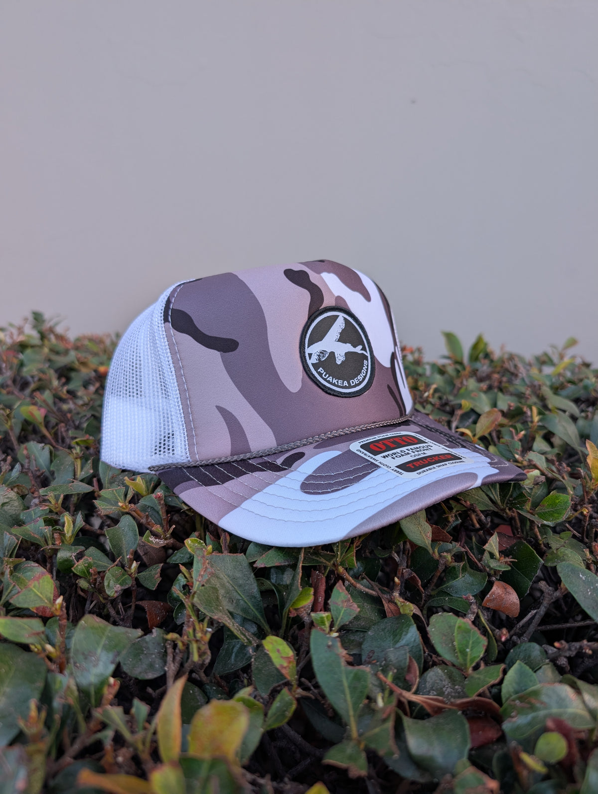 Puakea camo trucker hat with black-and-white logo patch, camouflage front and curved brim, and white mesh panels.
